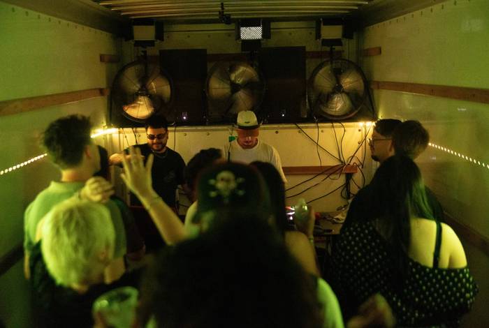 A crowd packed into the back of a box truck under green light, the DJ at the far end between three box fans